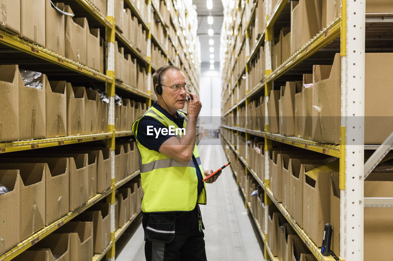 Senior male worker with digital tablet looking at packages on rack while talking through headset at distribution warehou