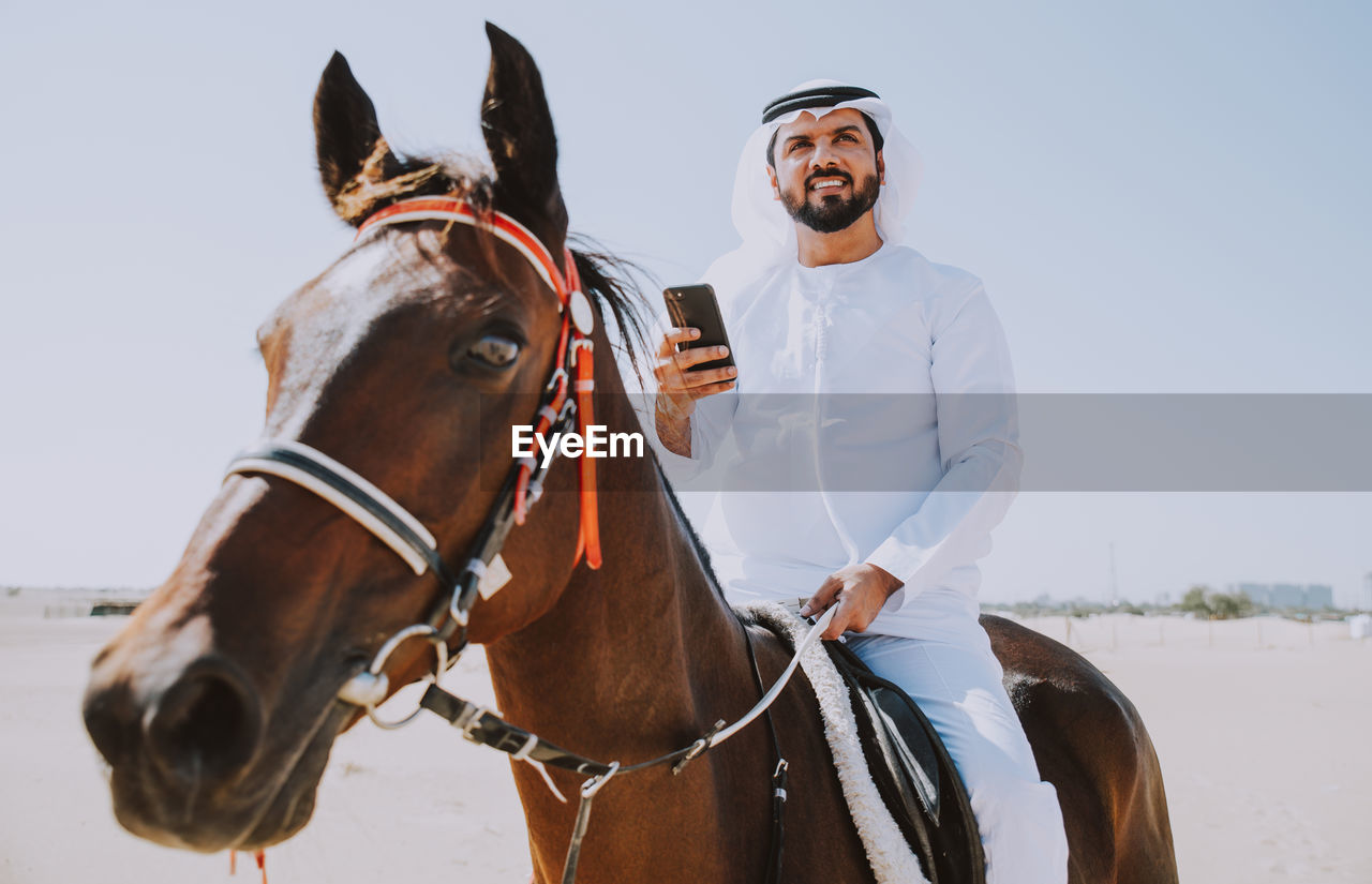 Man using mobile phone while horseback riding against sky