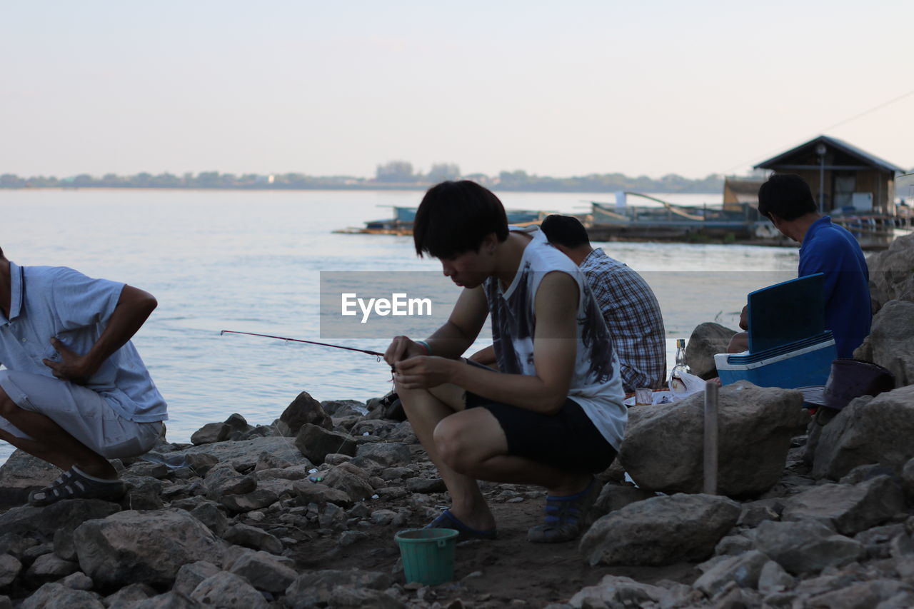 MEN SITTING ON ROCKS BY SEA AGAINST SKY