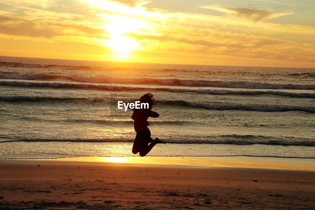 Silhouette of woman jumping at shore during sunset
