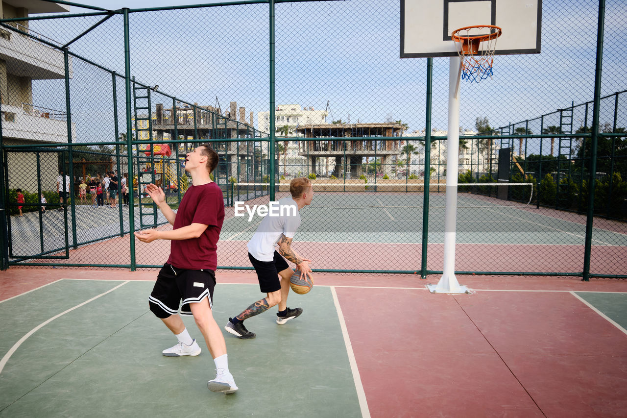 Two young adult men play basketball at outdoor court.