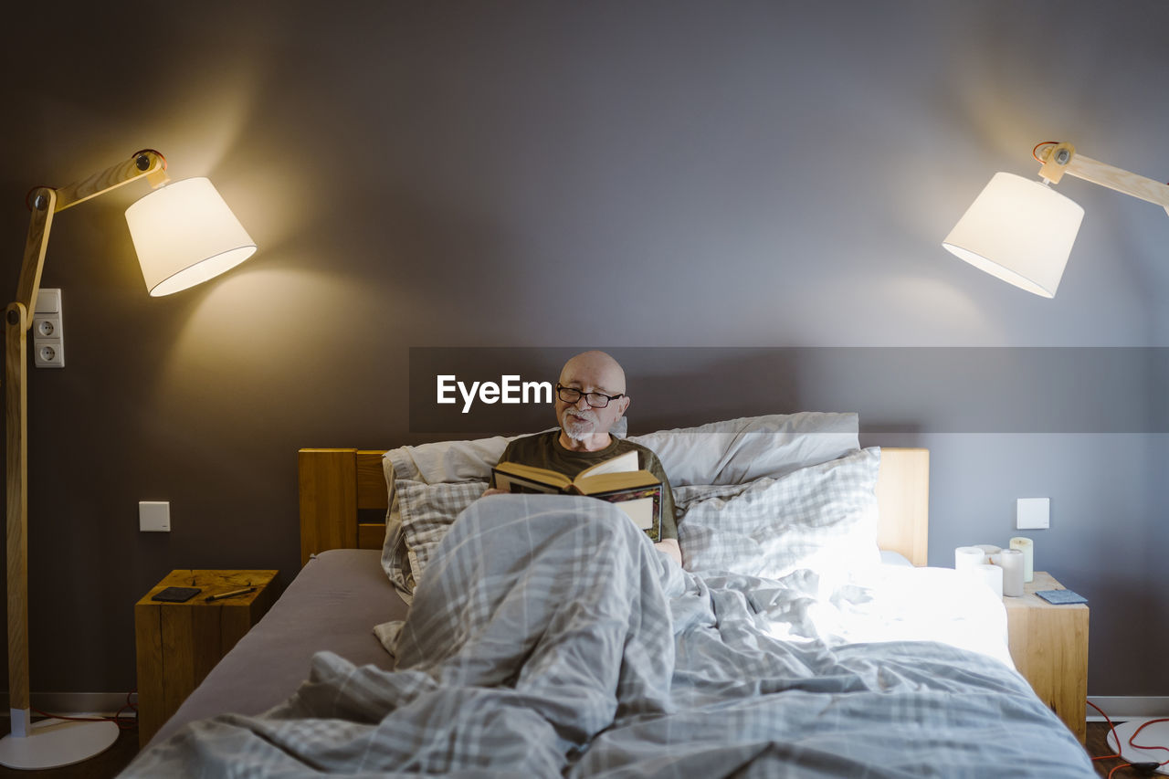 Senior man spending leisure time while reading book on bed at home