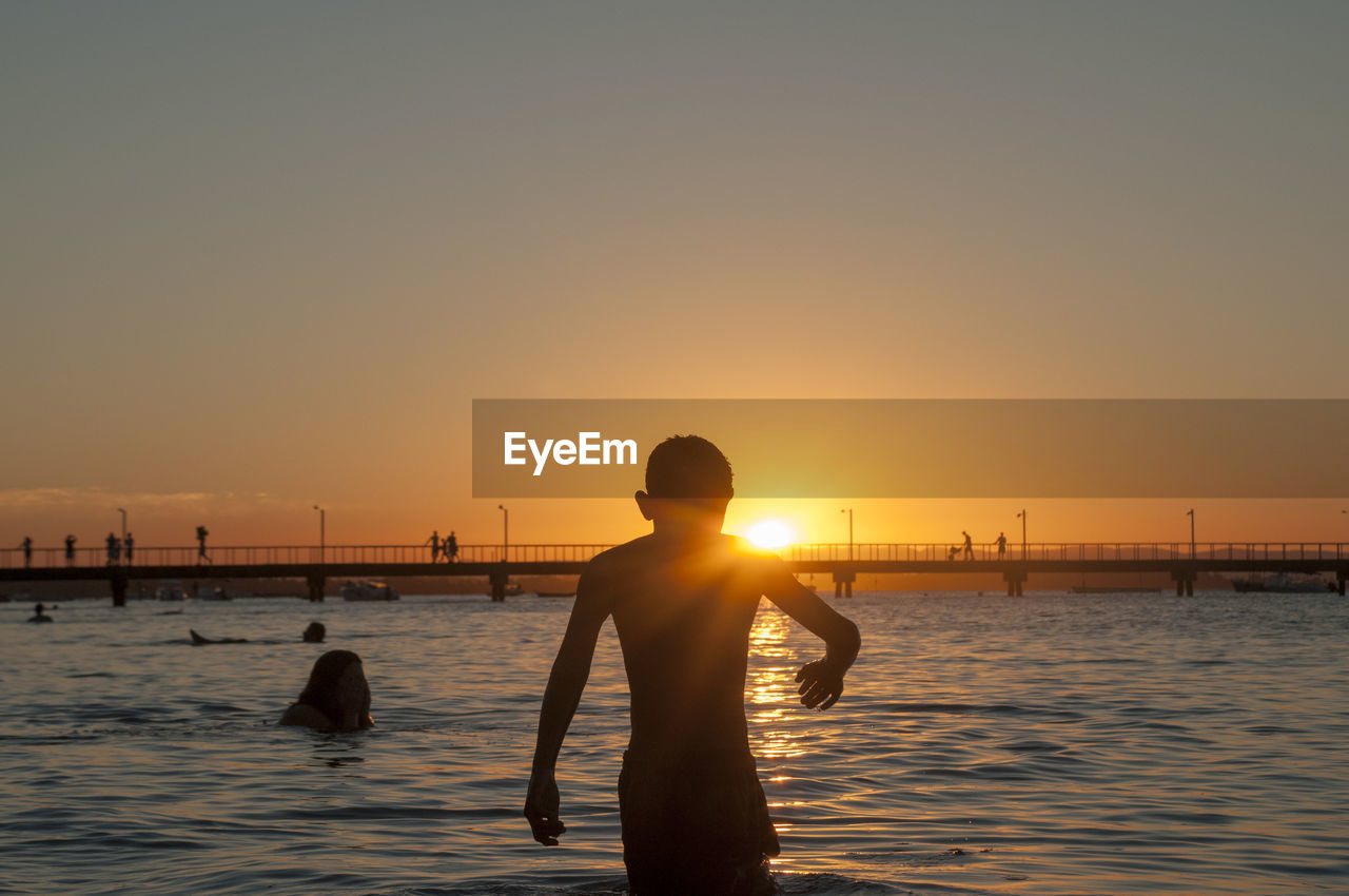 Silhouette boy standing in sea against sky during sunset