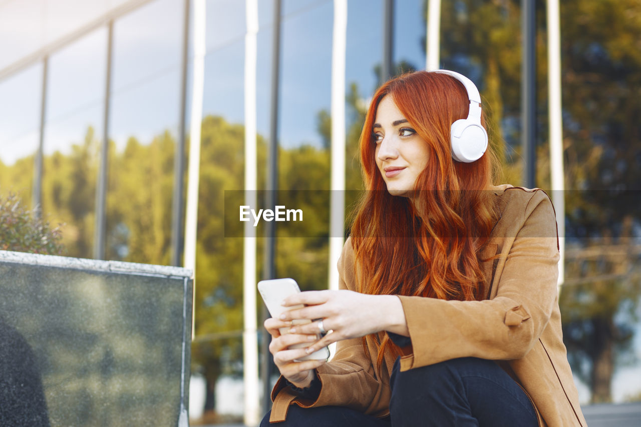 Young redhead woman sitting while listening to music with wireless headphones and smartphone,