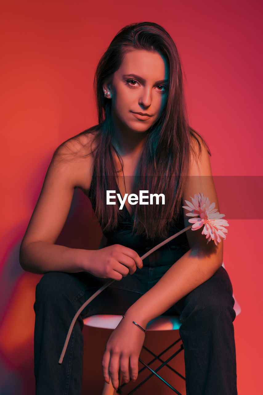 Portrait of young woman holding flower while sitting against red background