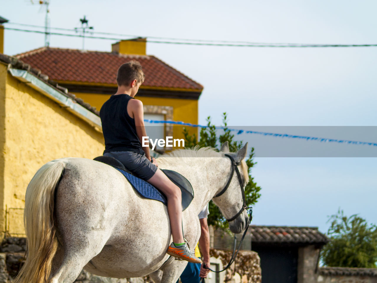 Boy riding horse by house against sky
