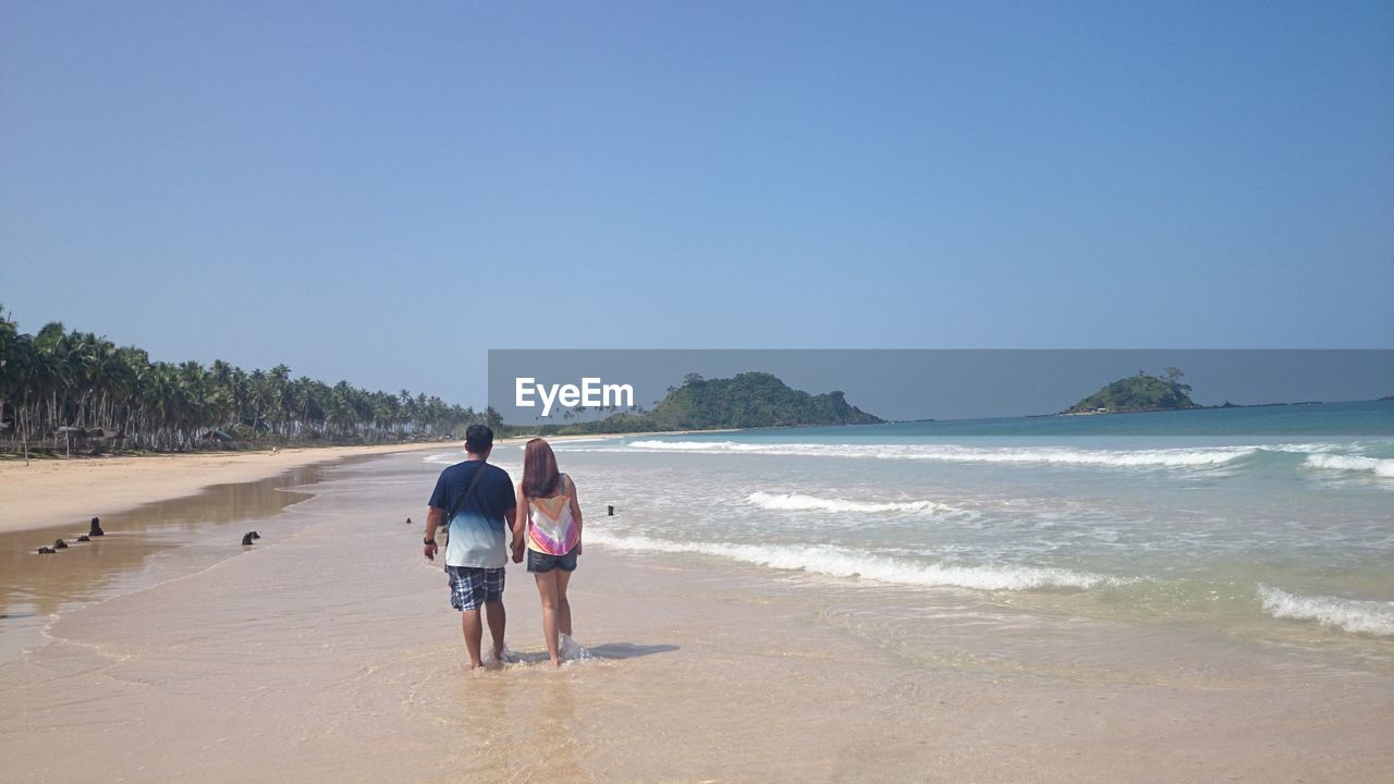 Rear view of couple walking on shore at beach against clear blue sky