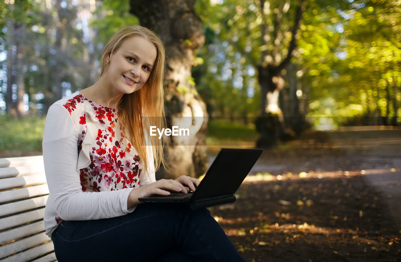 Portrait of smiling beautiful woman using laptop while sitting on bench at park