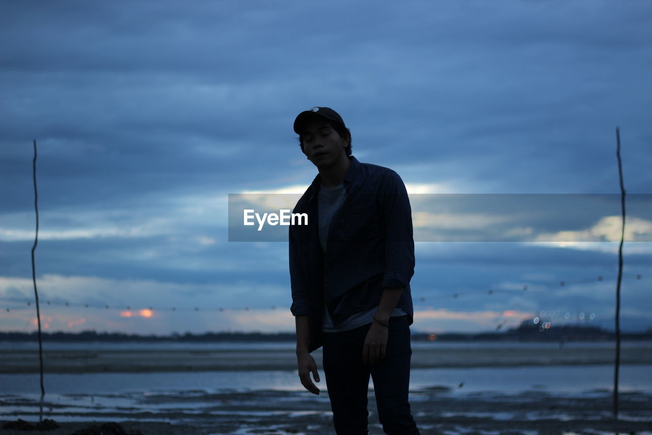 Young man standing at lakeshore against cloudy sky