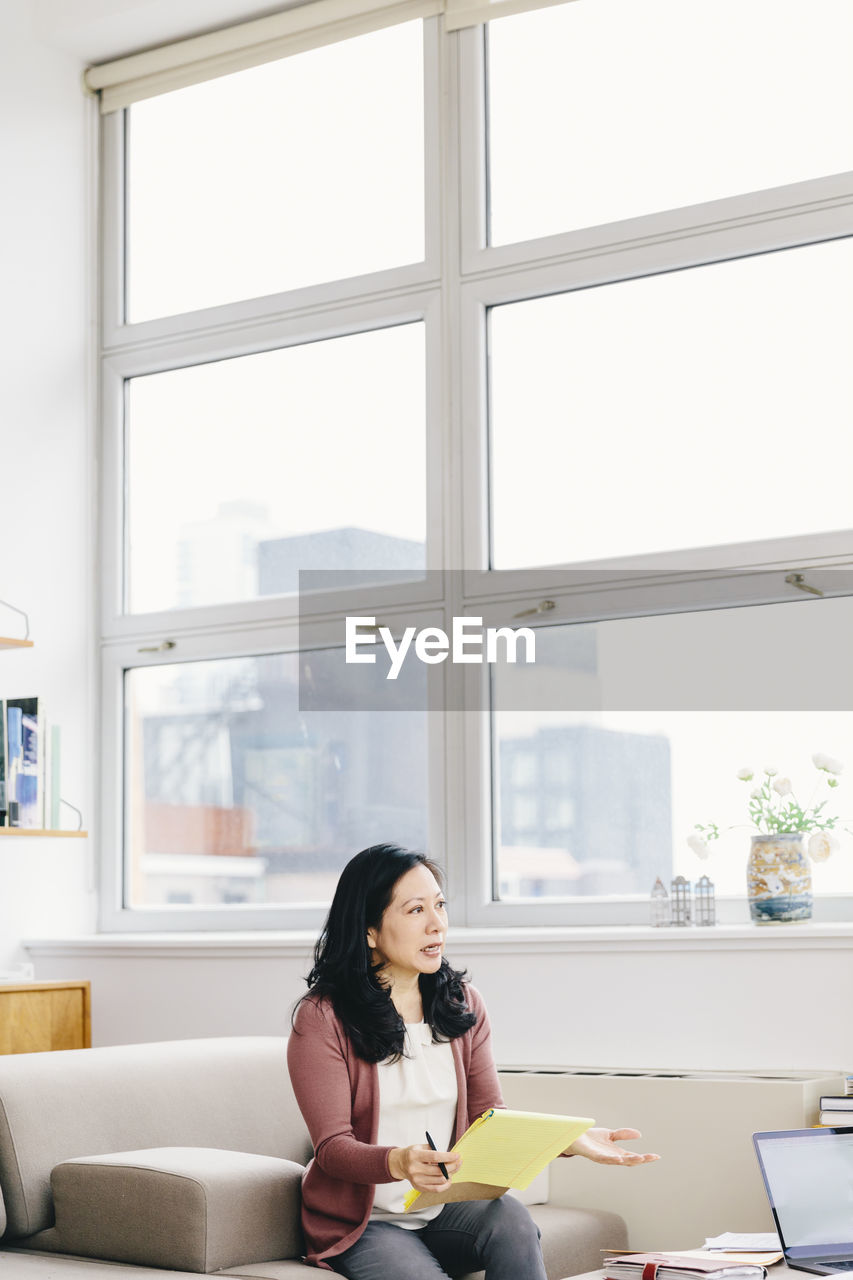 Businesswoman holding notepad while looking away against windows in office