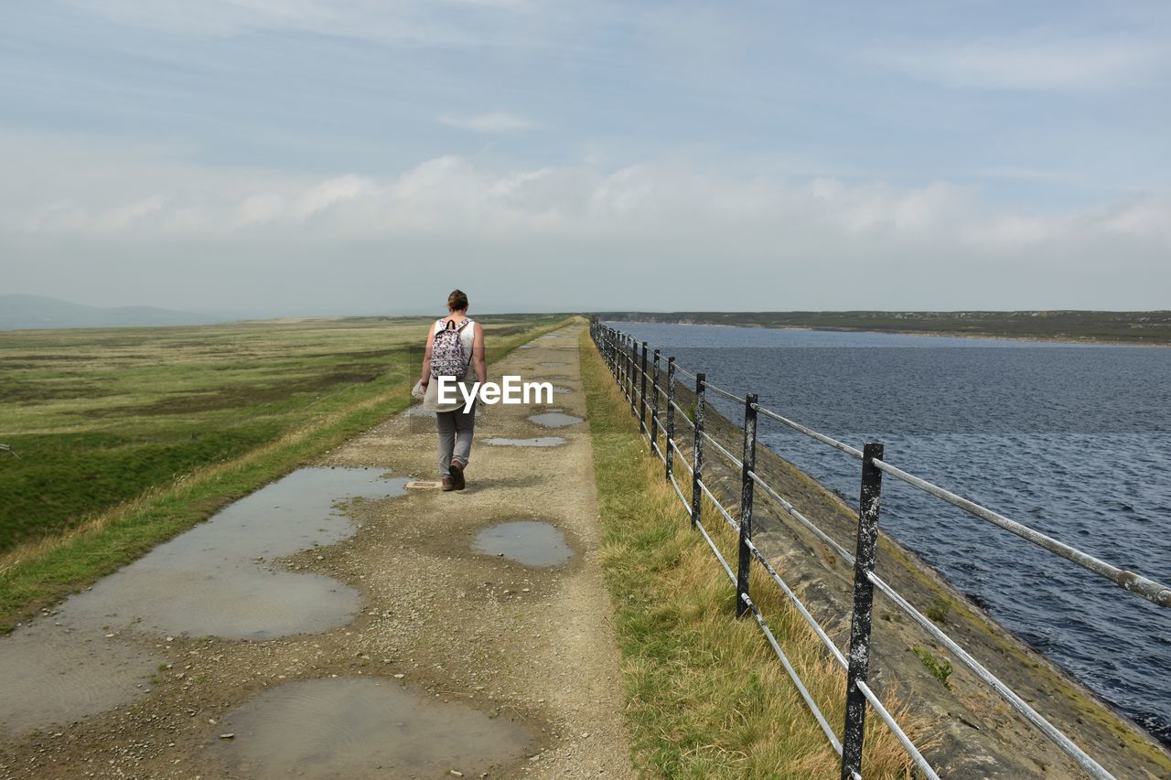 Rear view of woman walking on the dam of a reservoir in moorland against sky 