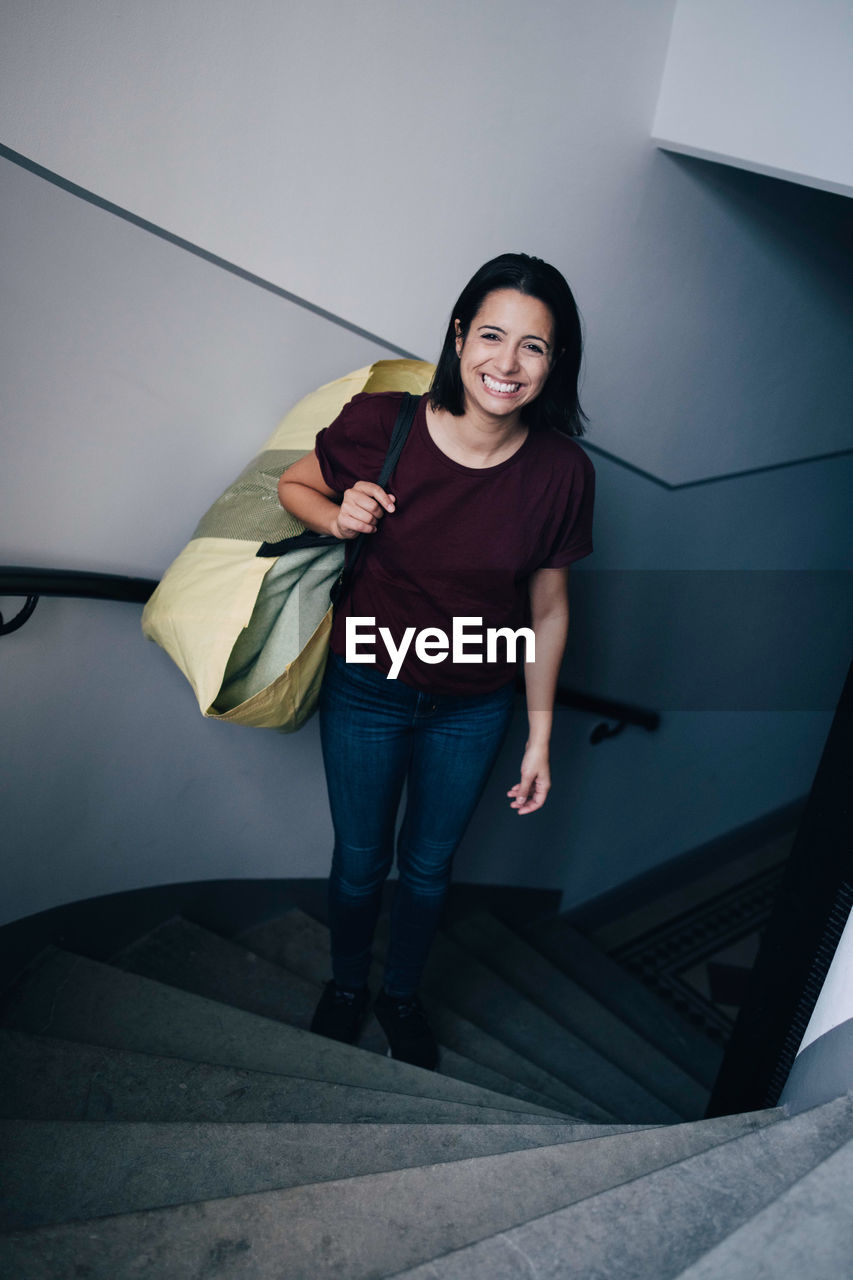 High angle portrait of smiling woman carrying bag while standing on steps