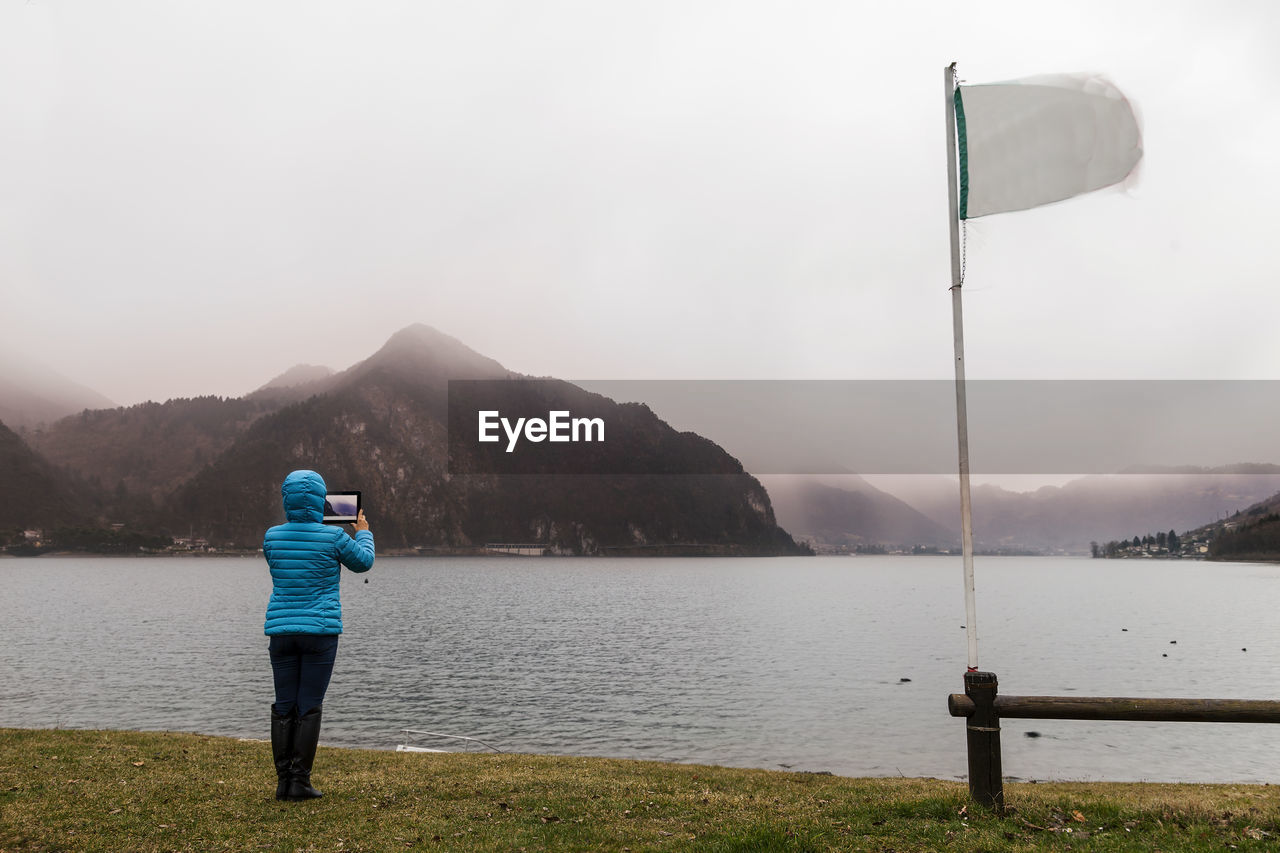 Rear view of woman photographing lake against sky