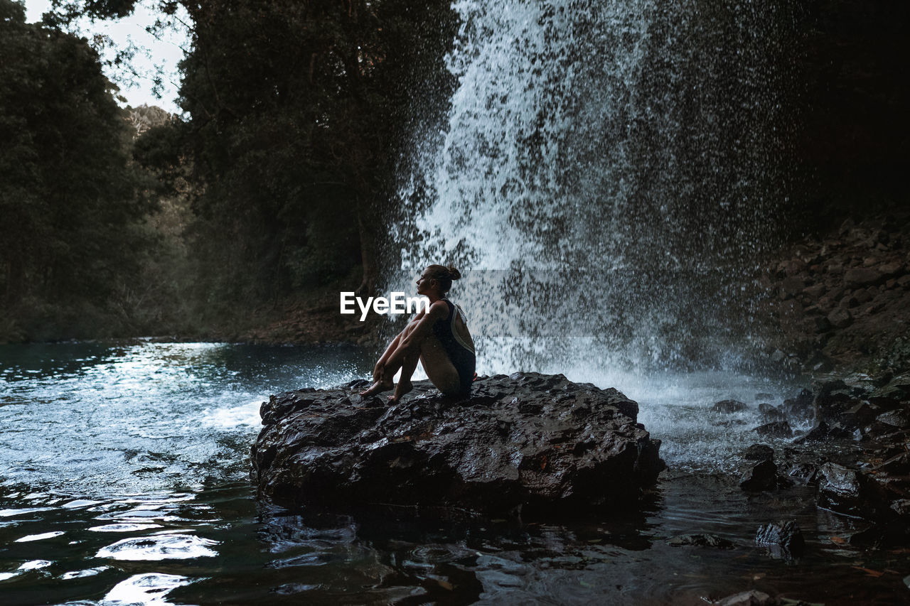 Side view of young woman sitting by waterfall on rock