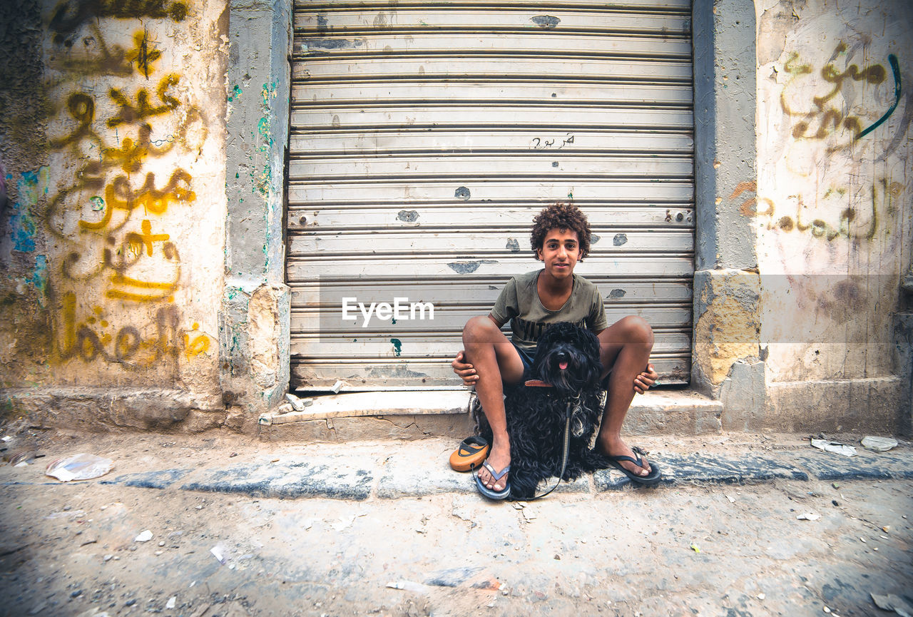 Portrait of young man sitting with dog against closed shutter in city