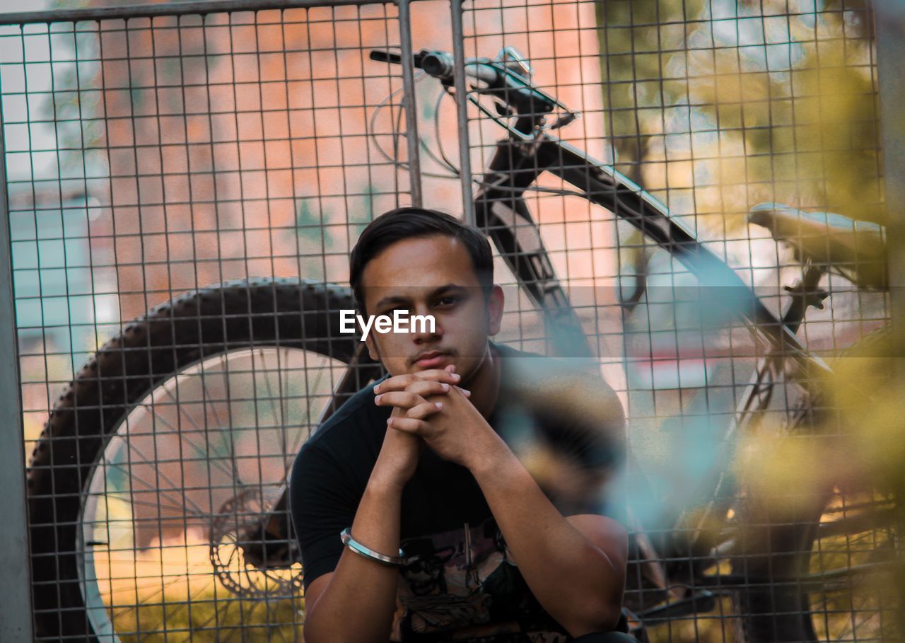 PORTRAIT OF YOUNG MAN LOOKING THROUGH FENCE AGAINST BLURRED BACKGROUND