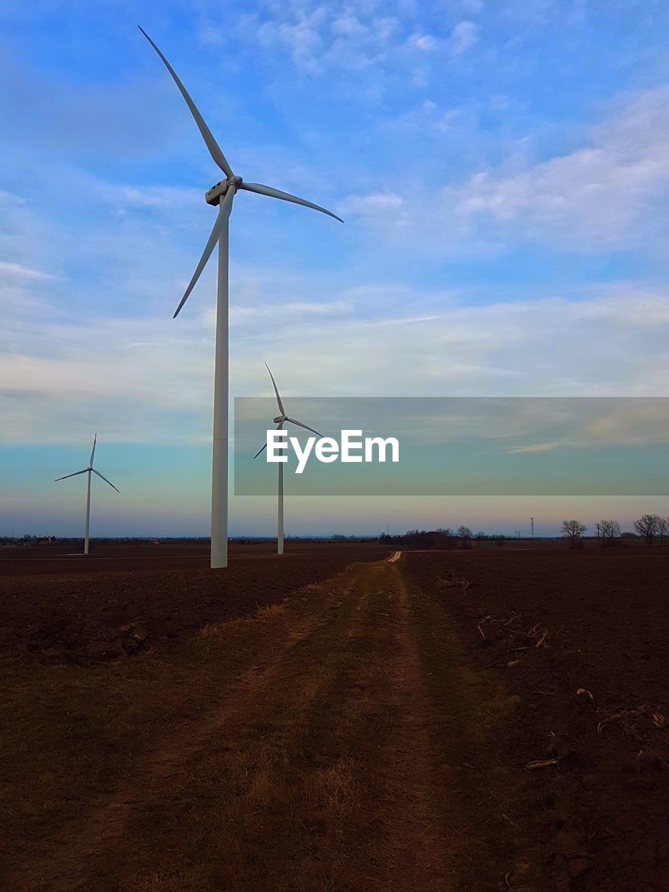 Dirt road by wind turbines on field against sky