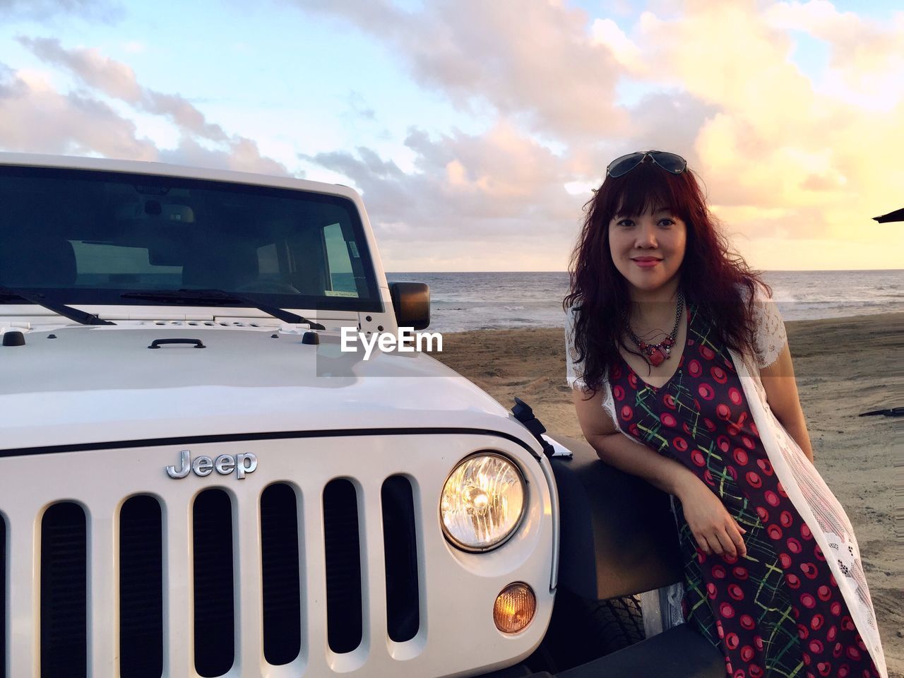 PORTRAIT OF A SMILING YOUNG WOMAN STANDING BY CAR AGAINST SKY