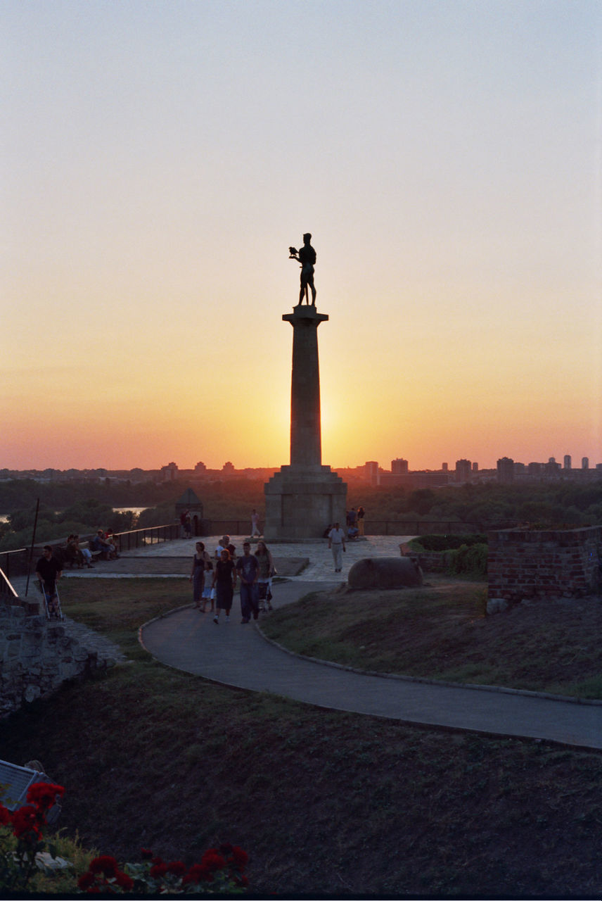 People with statue in park against sky during sunset