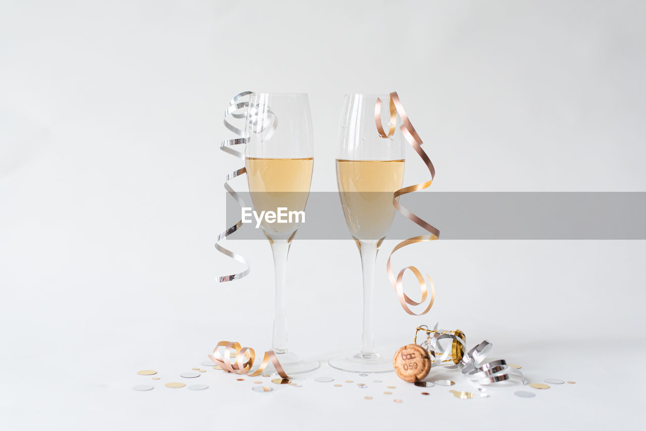 Two glasses of champagne, cork, ribbon and confetti on white backdrop.
