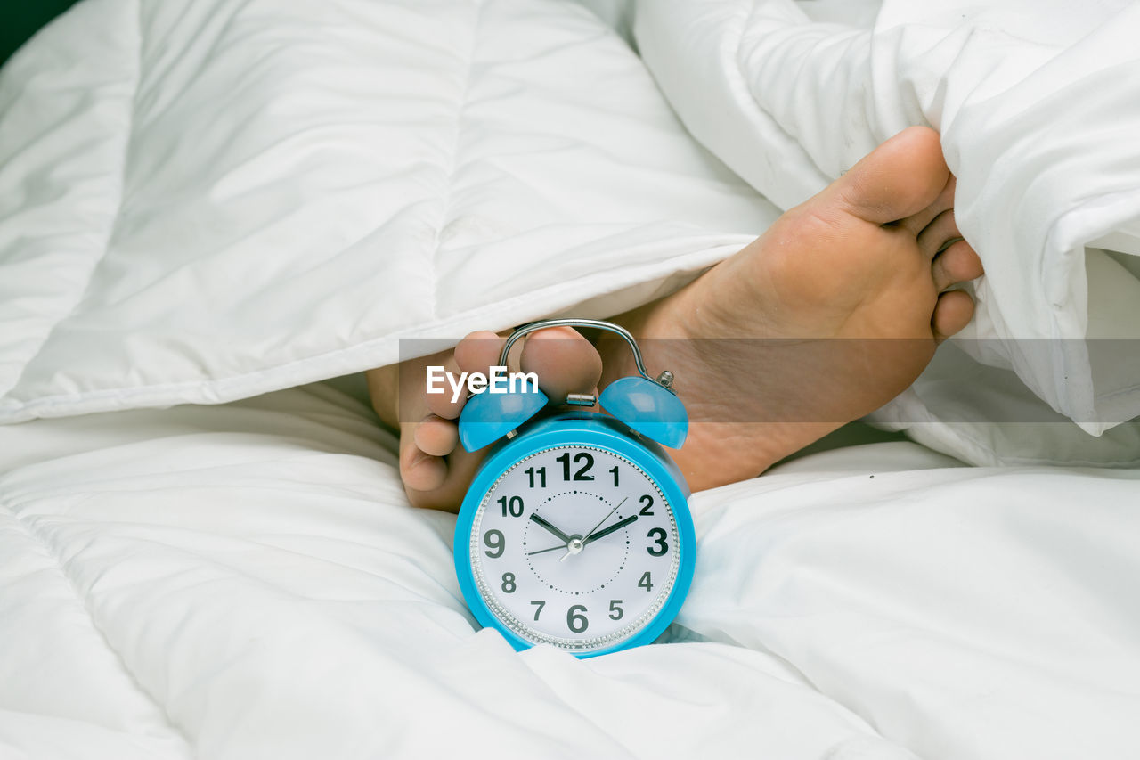 Alarm clock by low section of man sleeping on bed