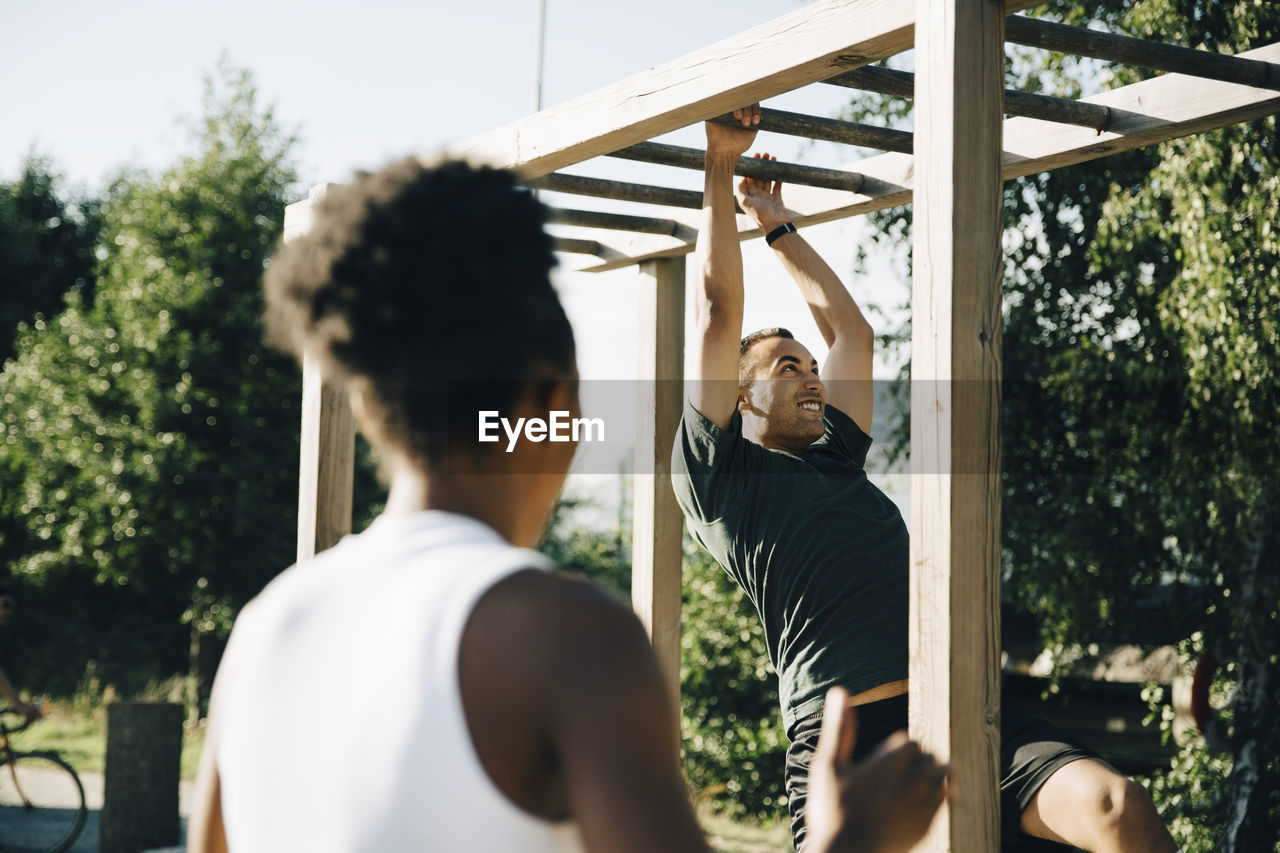 Male athlete hanging on monkey bar while sportswoman looking at him in park on sunny day