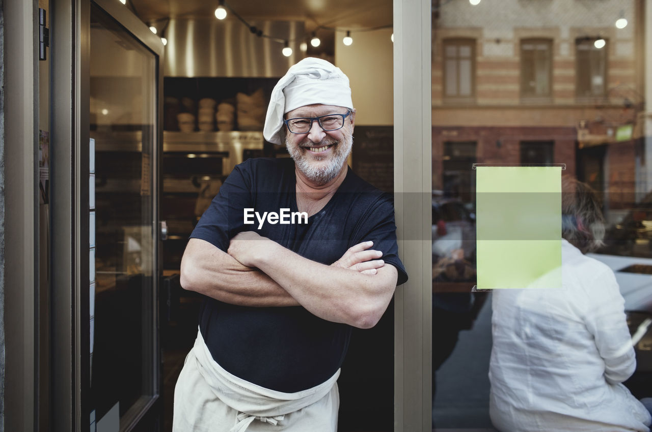 Smiling male baker standing with arms crossed at entrance of bakery