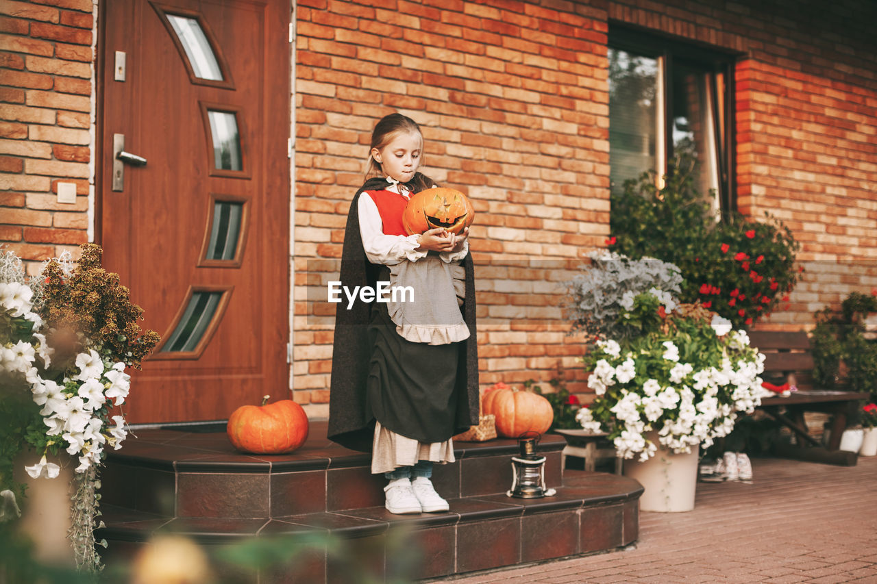 Full length of cute girl holding pumpkin standing against house