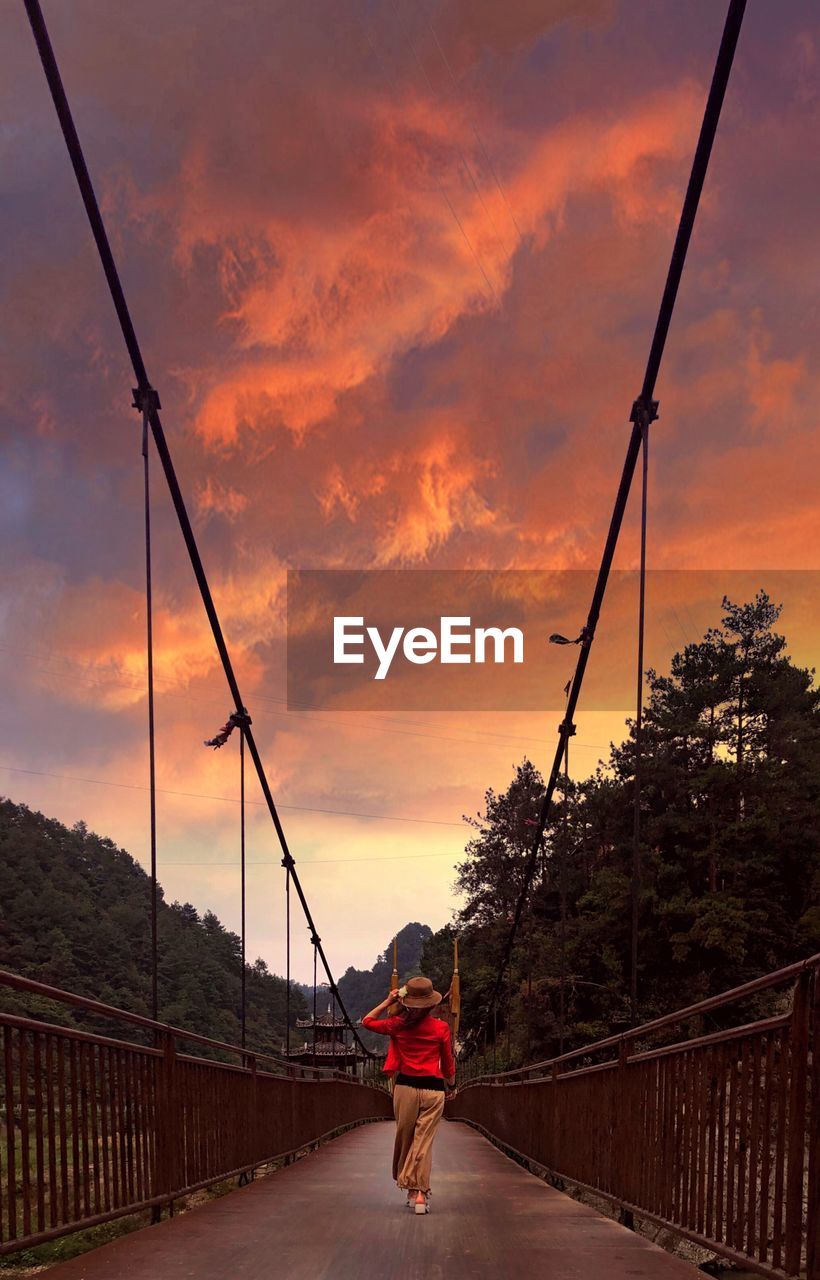 Rear view of woman walking on footbridge against sky during sunset