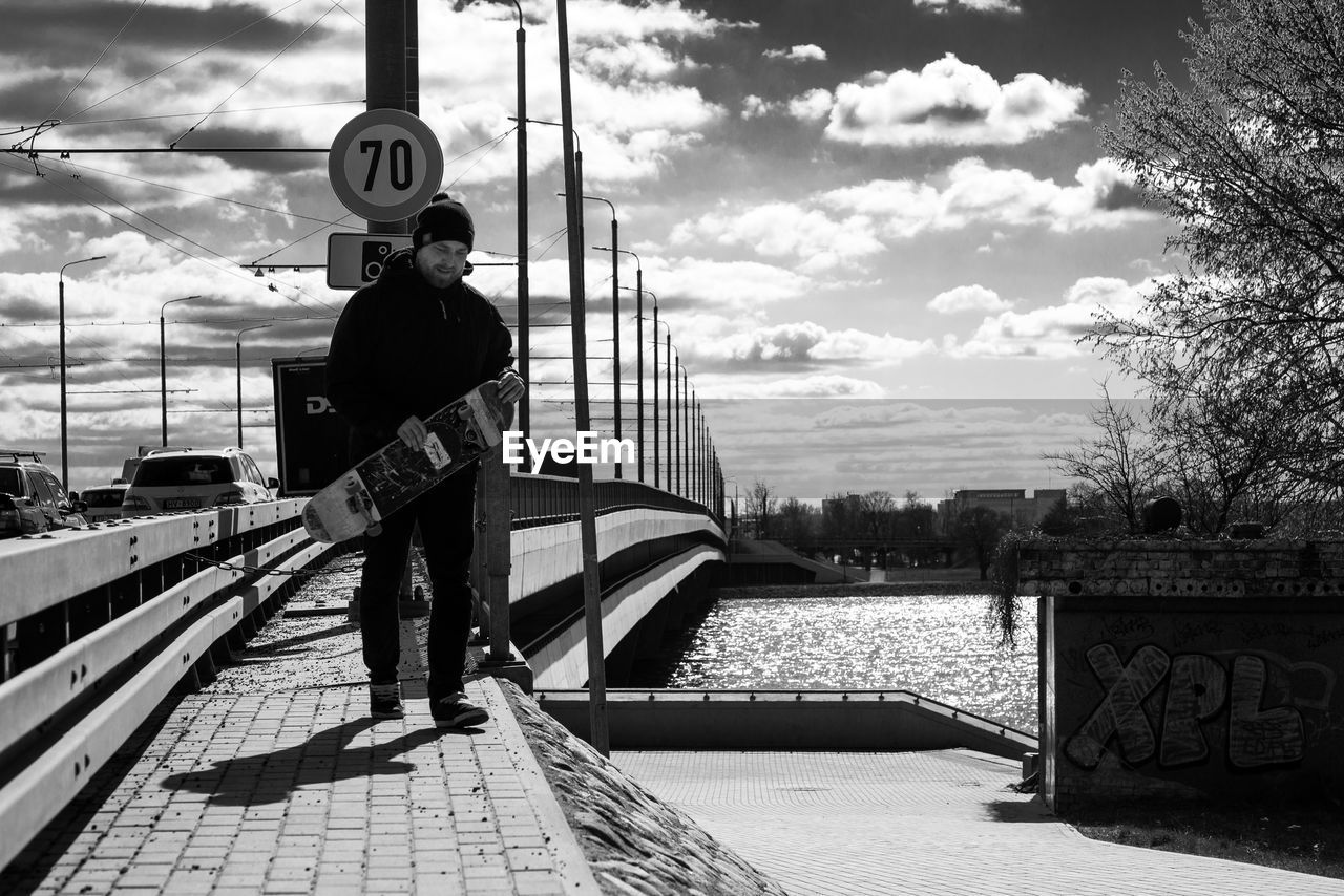 Man holding skateboard while standing on bridge against sky