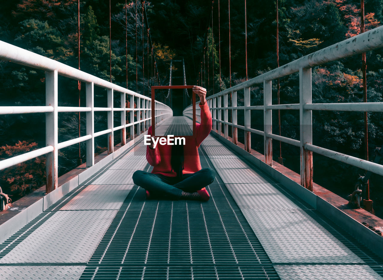 Woman covering face with mirror while sitting on footbridge