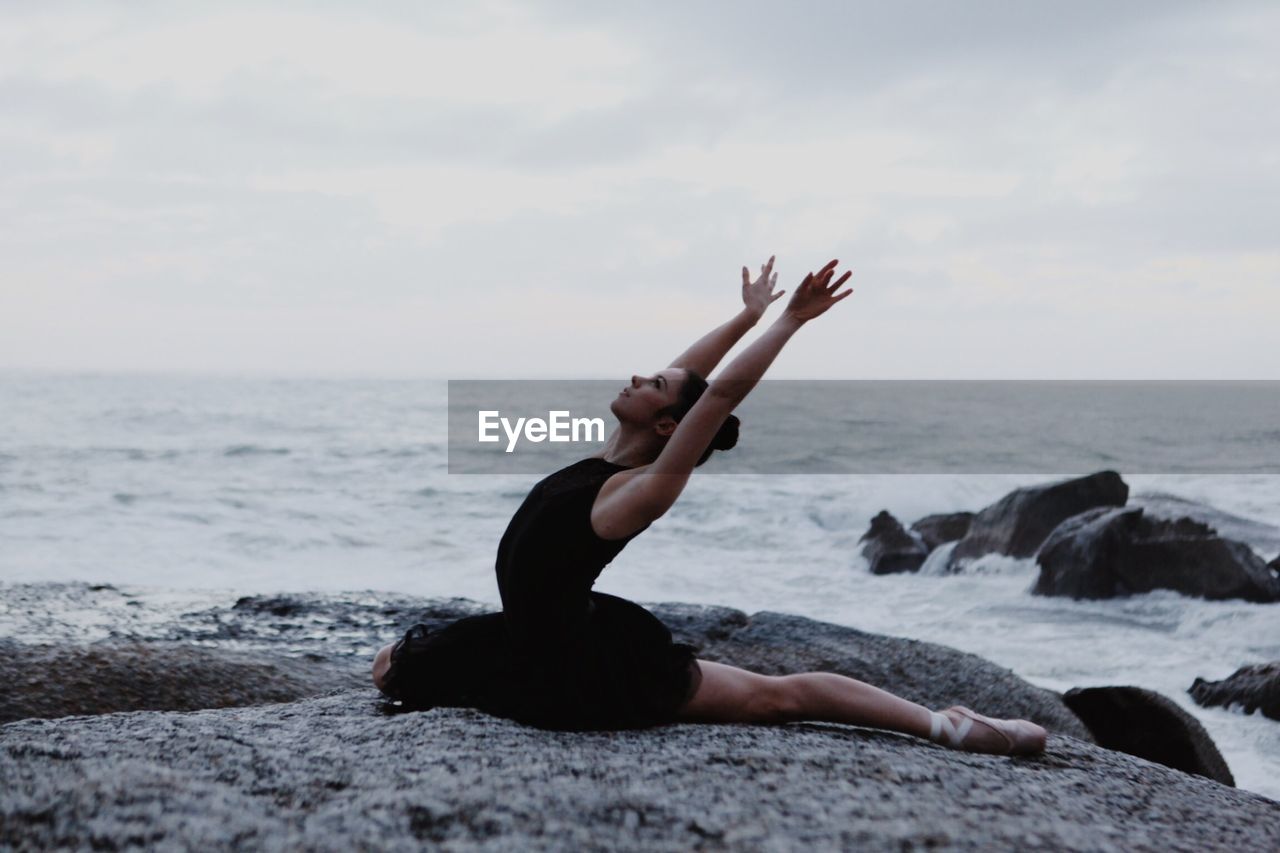 Side view of woman stretching on rocks in front of sea with arms raised