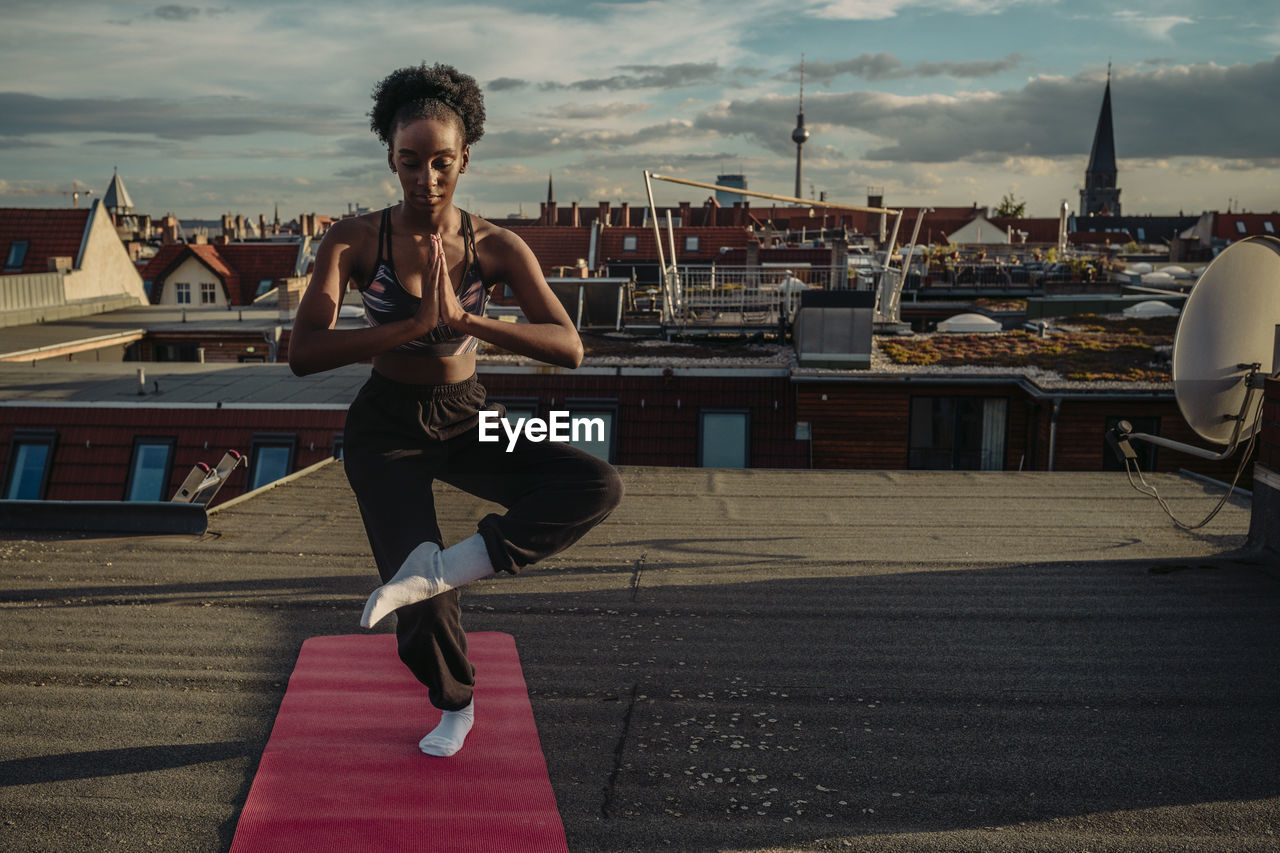 Young female standing on one leg while exercising on rooftop during sunrise