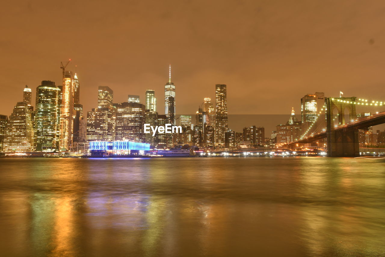 Illuminated modern buildings by river against sky at night