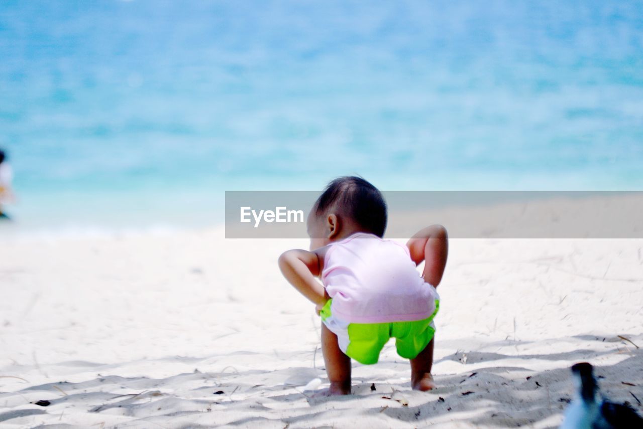 Rear view of baby boy crouching on sand at beach against sky