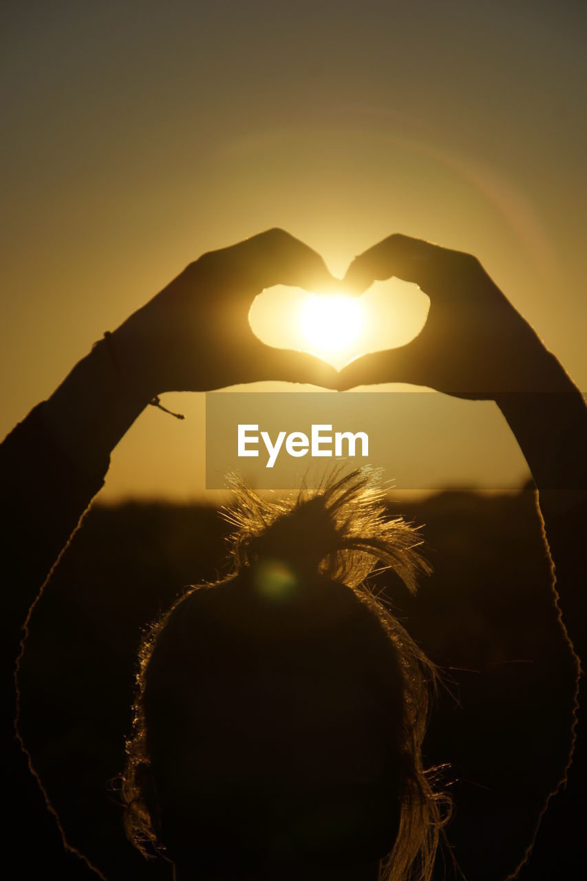 Close-up of woman making heart shape against sky during sunset