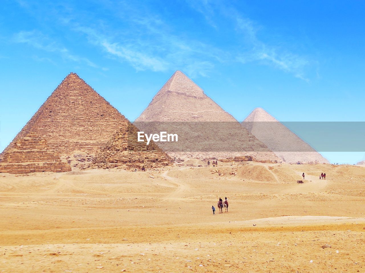 pyramid, travel destinations, desert, ancient, history, the past, travel, architecture, ancient civilization, sky, nature, camel, climate, domestic animals, land, tourism, scenics - nature, sand, city, shape, built structure, arid climate, landscape, mammal, environment, triangle shape, monument, geometric shape, animal, animal themes, tranquil scene, cloud, blue, day, building exterior, clear sky, outdoors, sunlight, archaeology, distant, tranquility, beauty in nature, sunny, old ruin, ancient history, memorial, pyramid shape, riding, on the move, tomb, group of animals, transportation, pet, adult