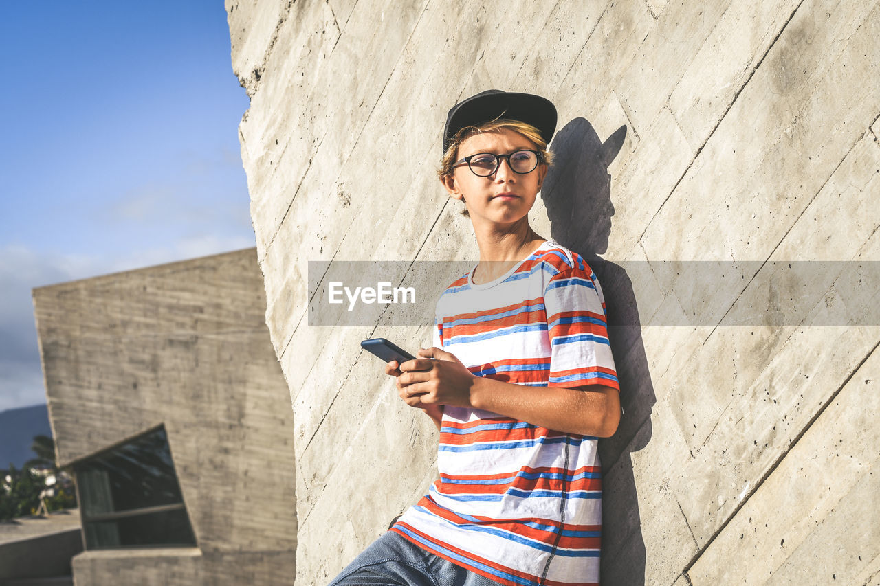 Boy holding phone while standing outside building on sunny day