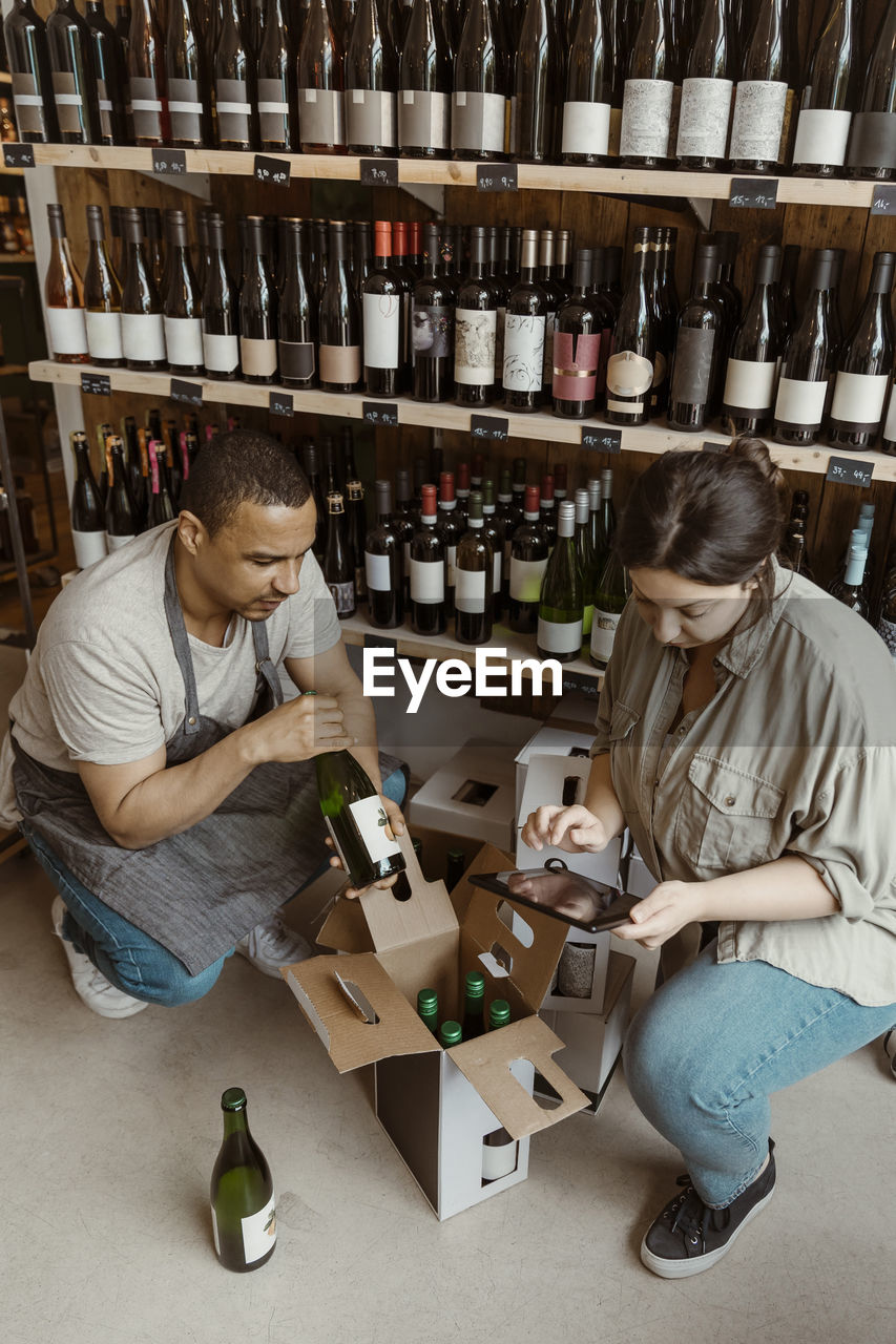 Male and female owners checking inventory of bottles while crouching in wine store