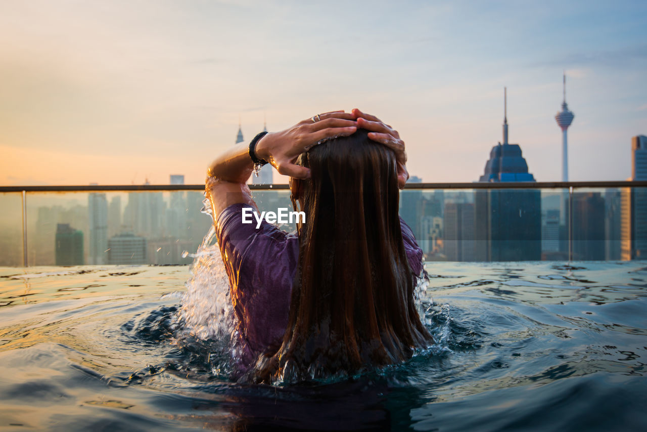 Rear view of woman swimming in infinity pool against city