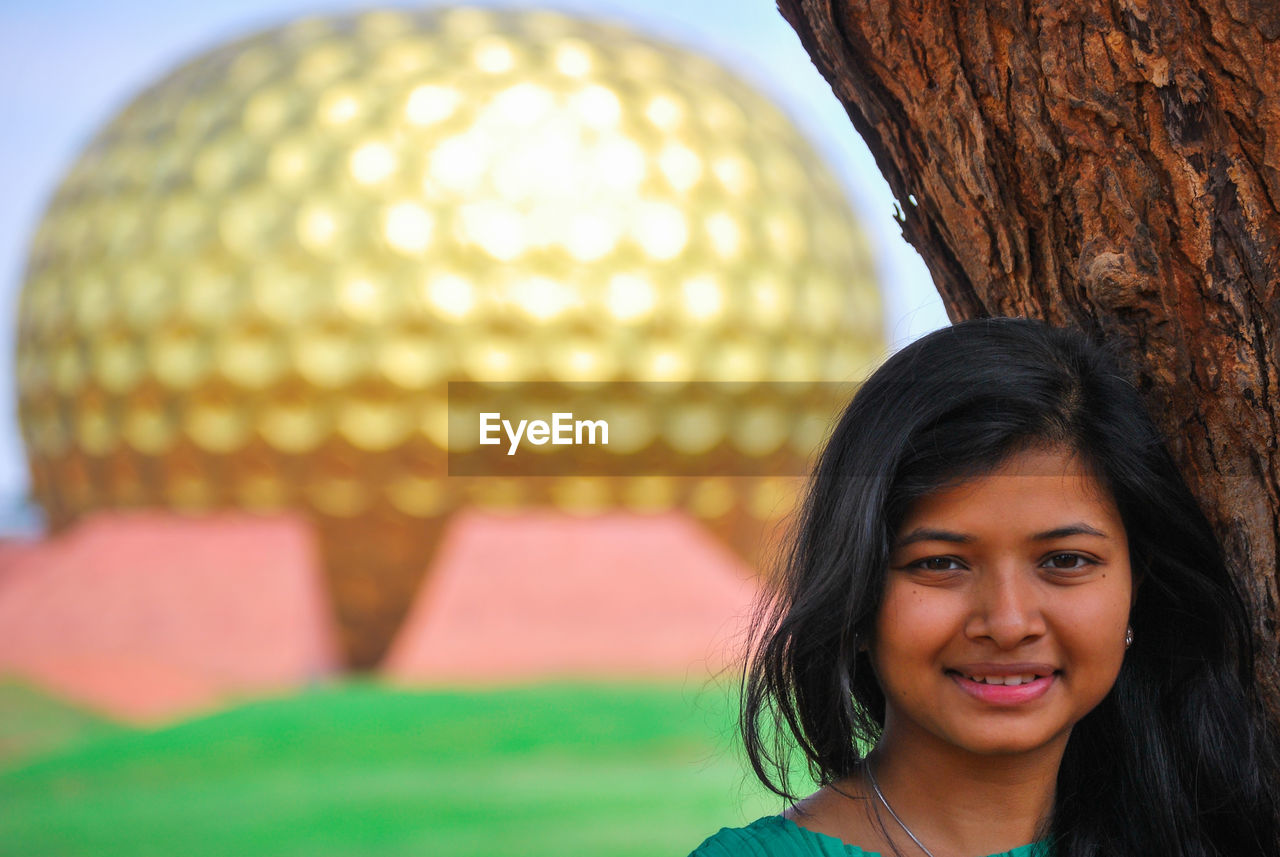 Portrait of smiling young woman standing by tree trunk