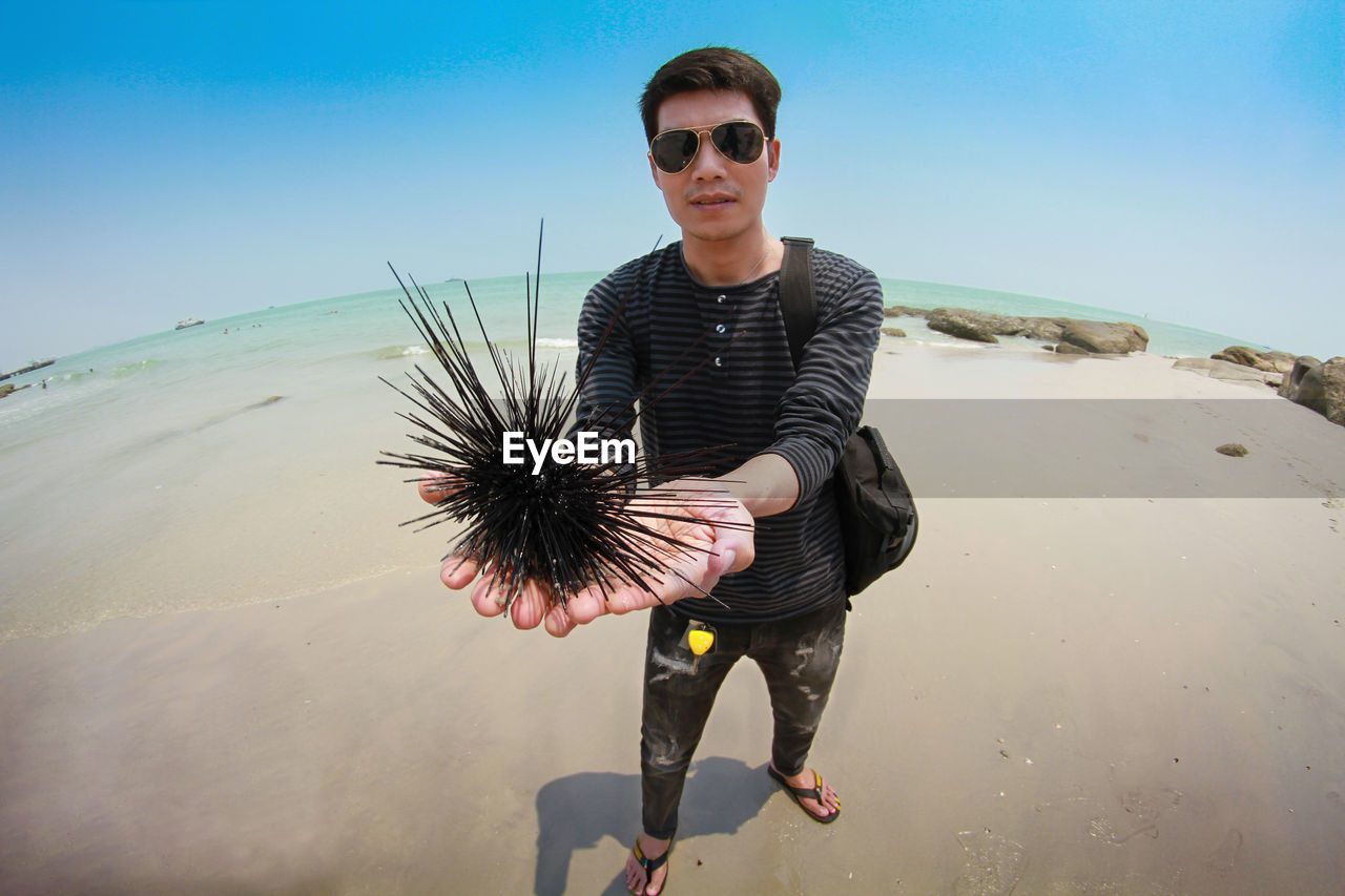 Portrait of young man holding urchin at beach against sky