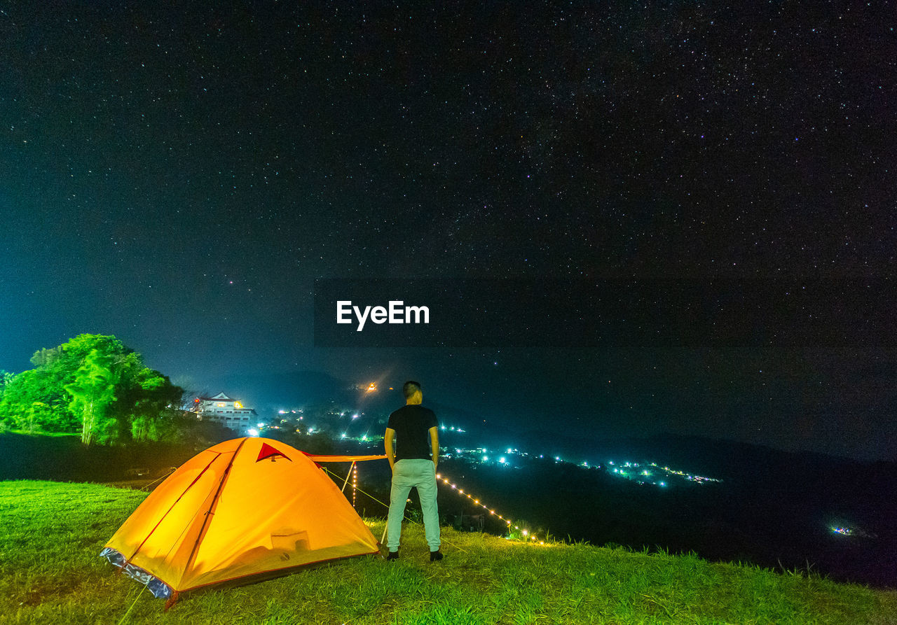 Rear view of man standing by illuminated tent against sky at night