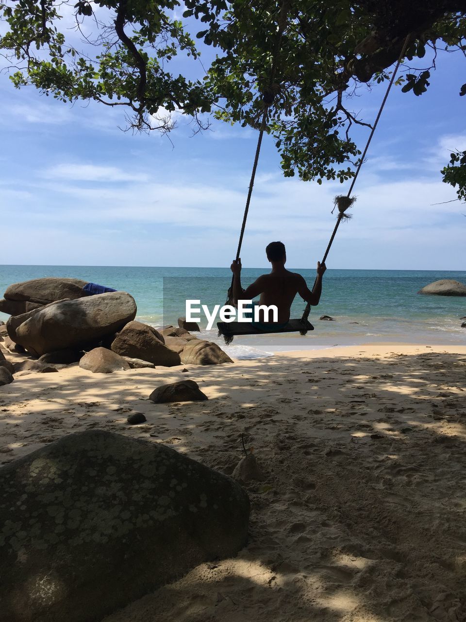 Shirtless man swinging at beach against sky