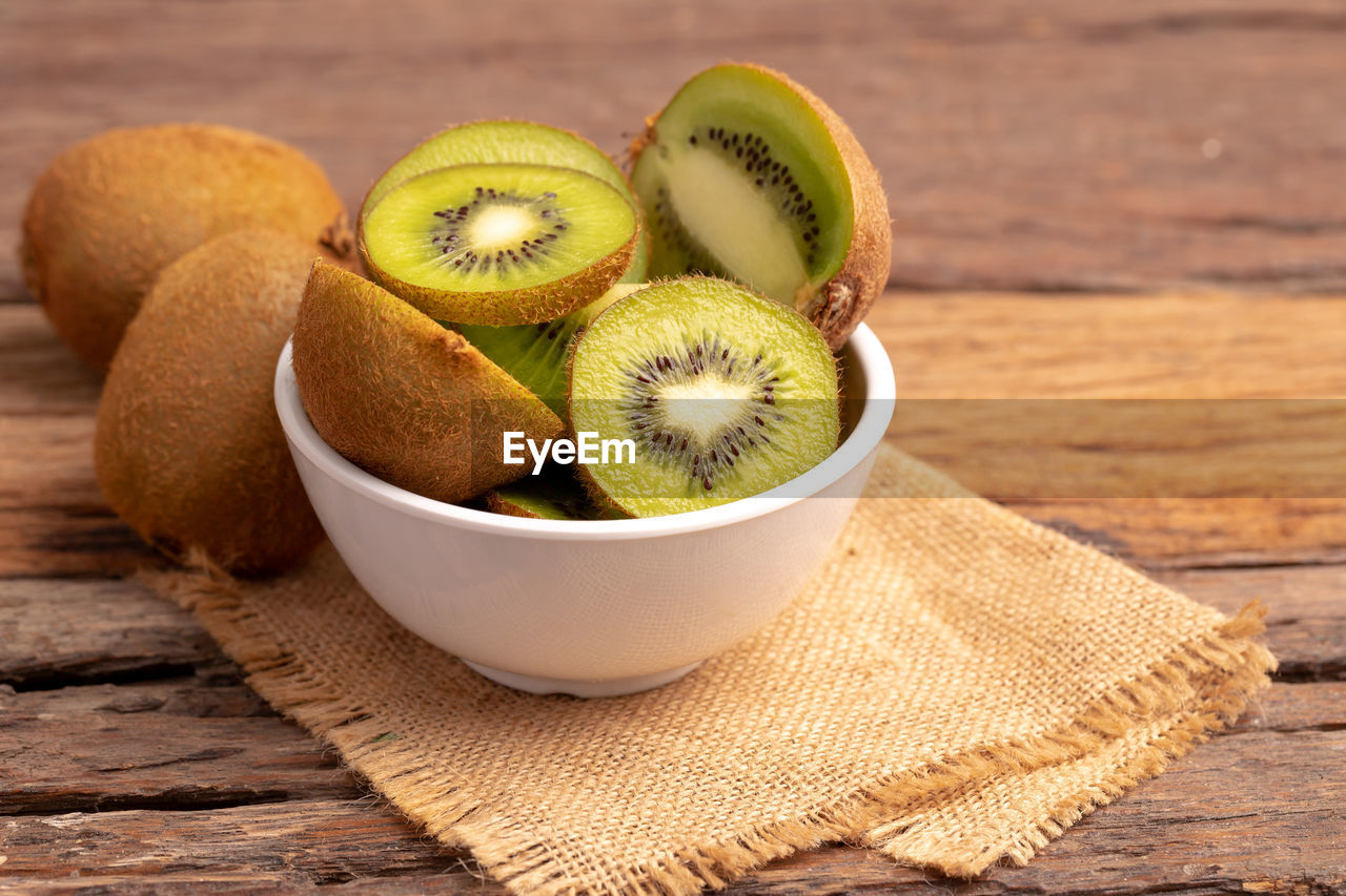 Slices of fresh kiwis in a white bowl on the brown sack placed on the old wooden table