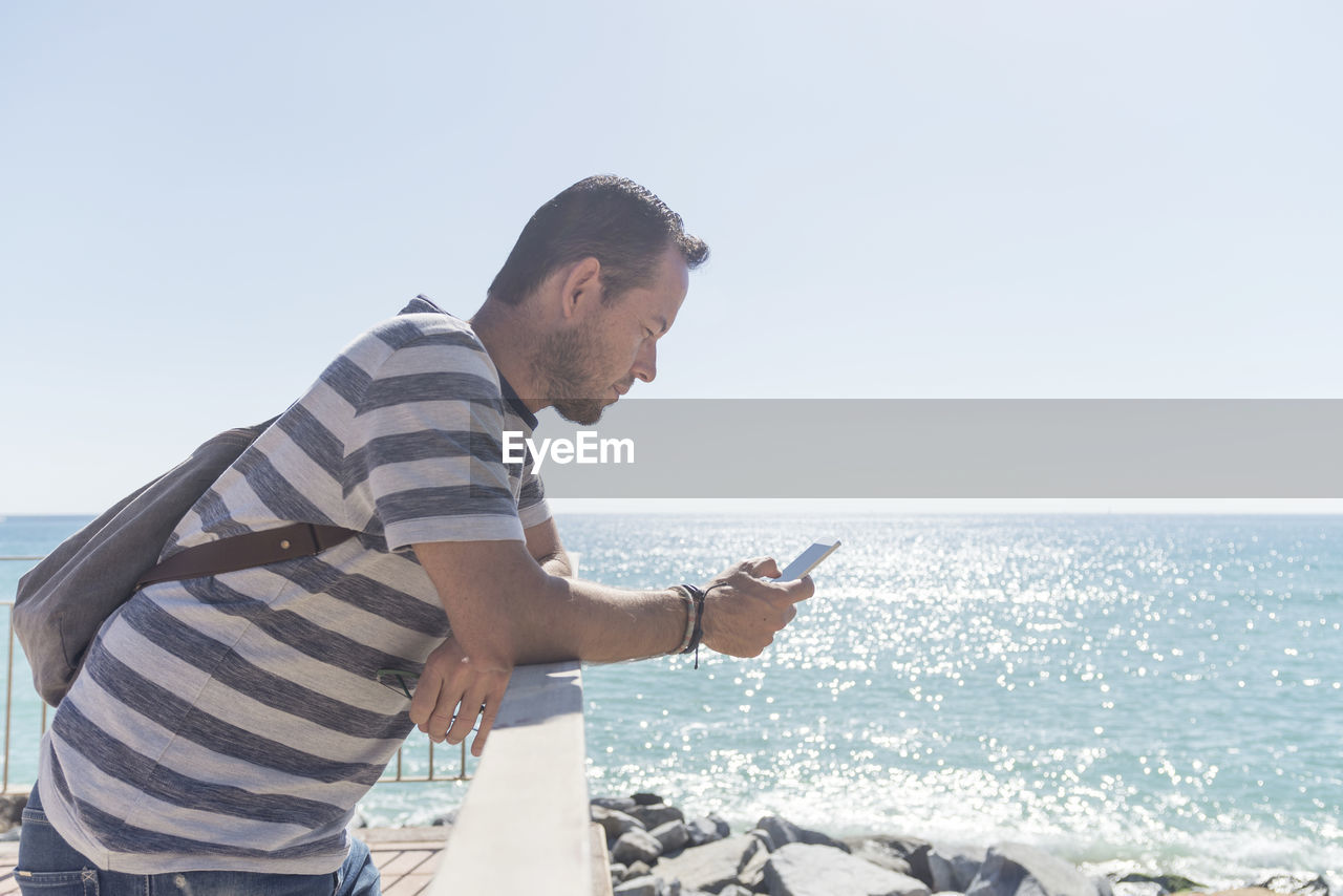 Side view of man using phone while standing against sea on sunny day