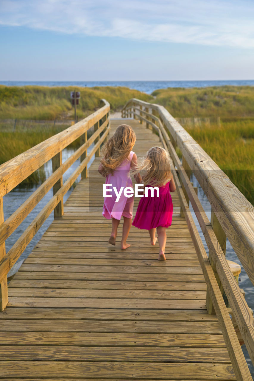 Little girls from behind running on bridge to beach in pink dresses