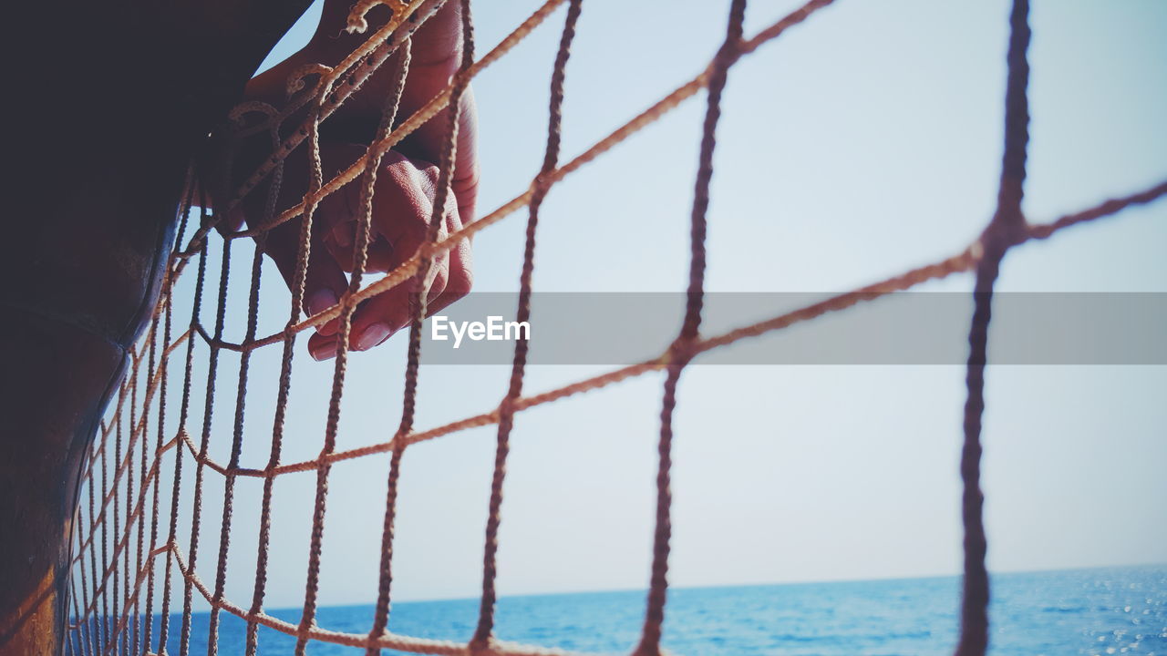 Cropped image of hands on net by sea against clear sky