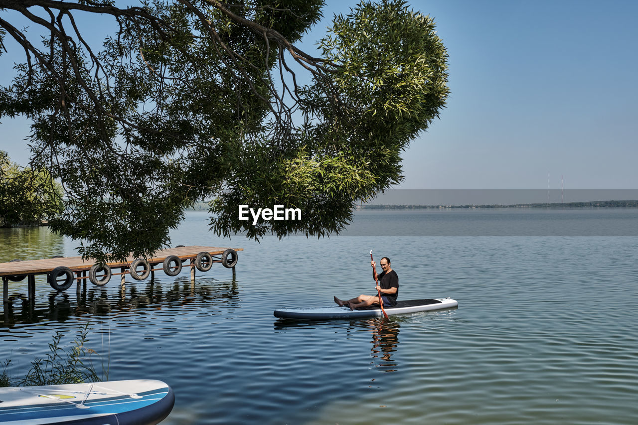 Asian older man sitting on sup board, rowing with oar on calm lake, floating to pier.