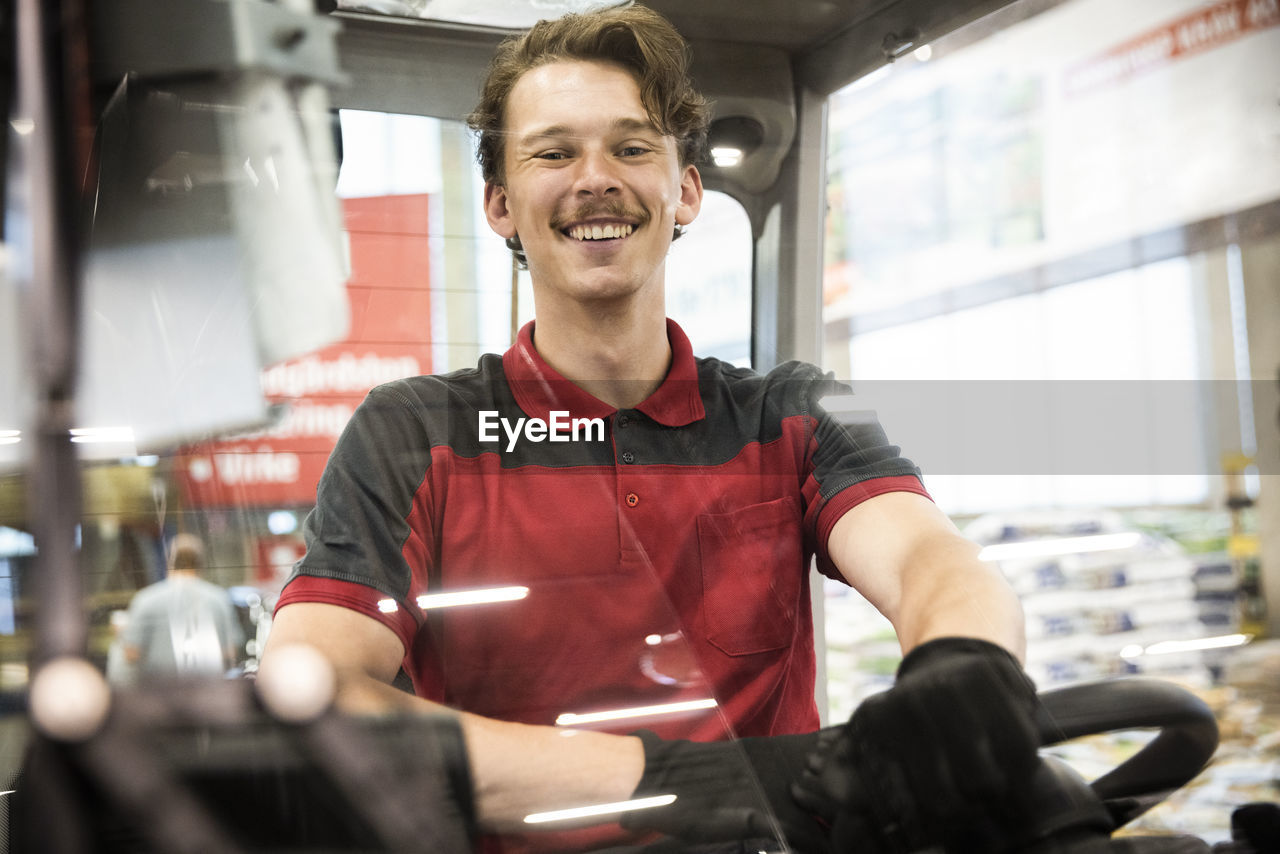 Portrait of happy male warehouse worker sitting in forklift seen through windshield